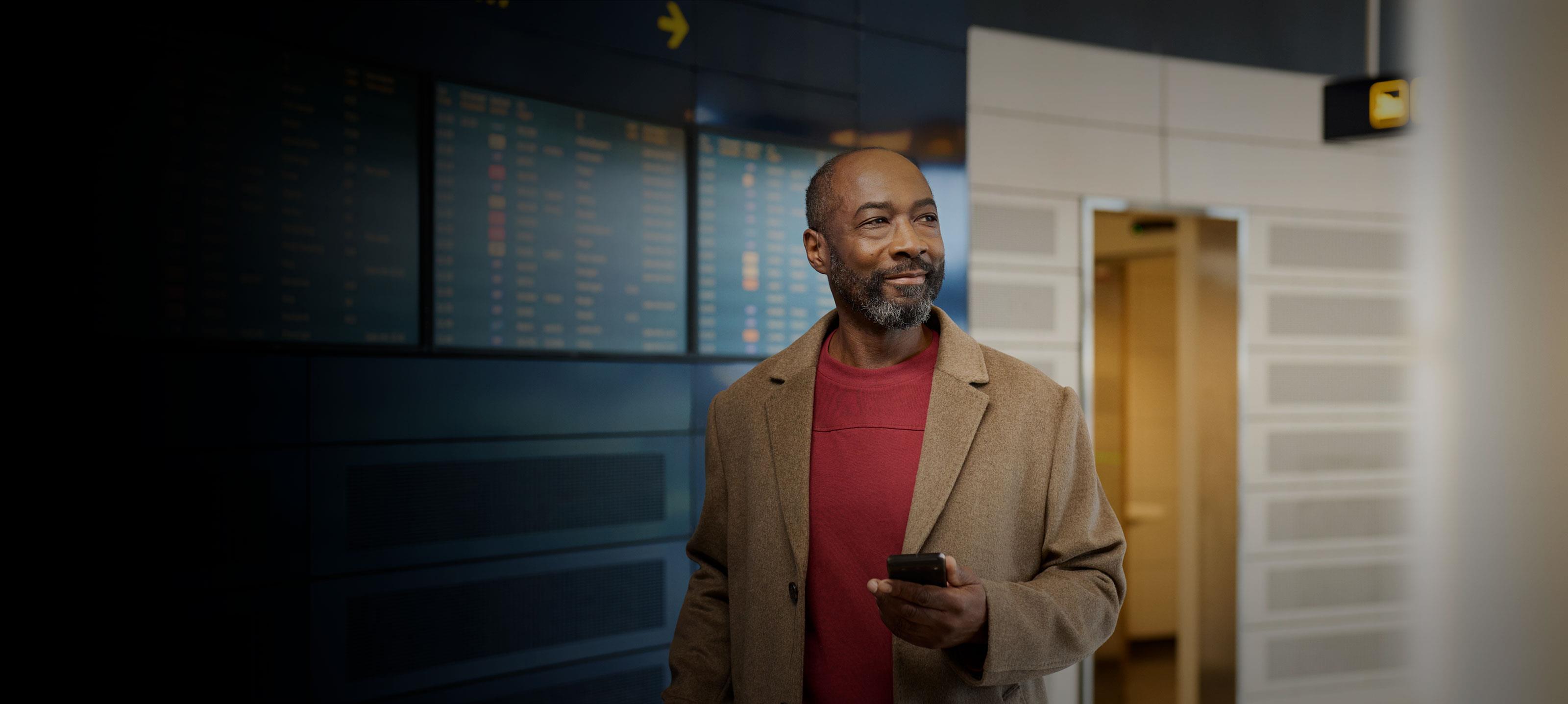 Man in a light brown coat and red sweater holding a mobile device in his hand in the airport